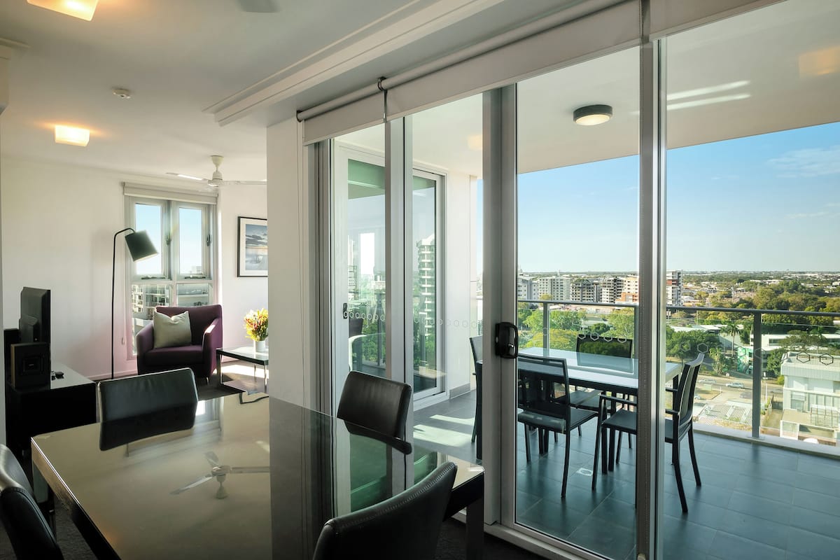 A modern living room with a glass table, chairs, and a sofa. There is a balcony with a view of the city.
