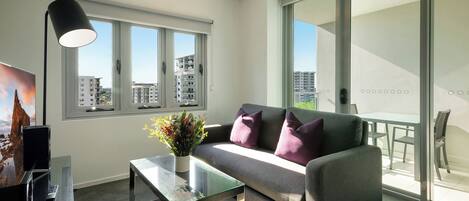 Un salon moderne avec un canapé, une table basse en verre, une lampe et une vue sur la ville grâce à de grandes fenêtres.