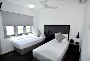 Una habitación de hotel con dos camas individuales, un ventilador de techo, una ventana con persianas y una mesita de noche con lámpara y teléfono.