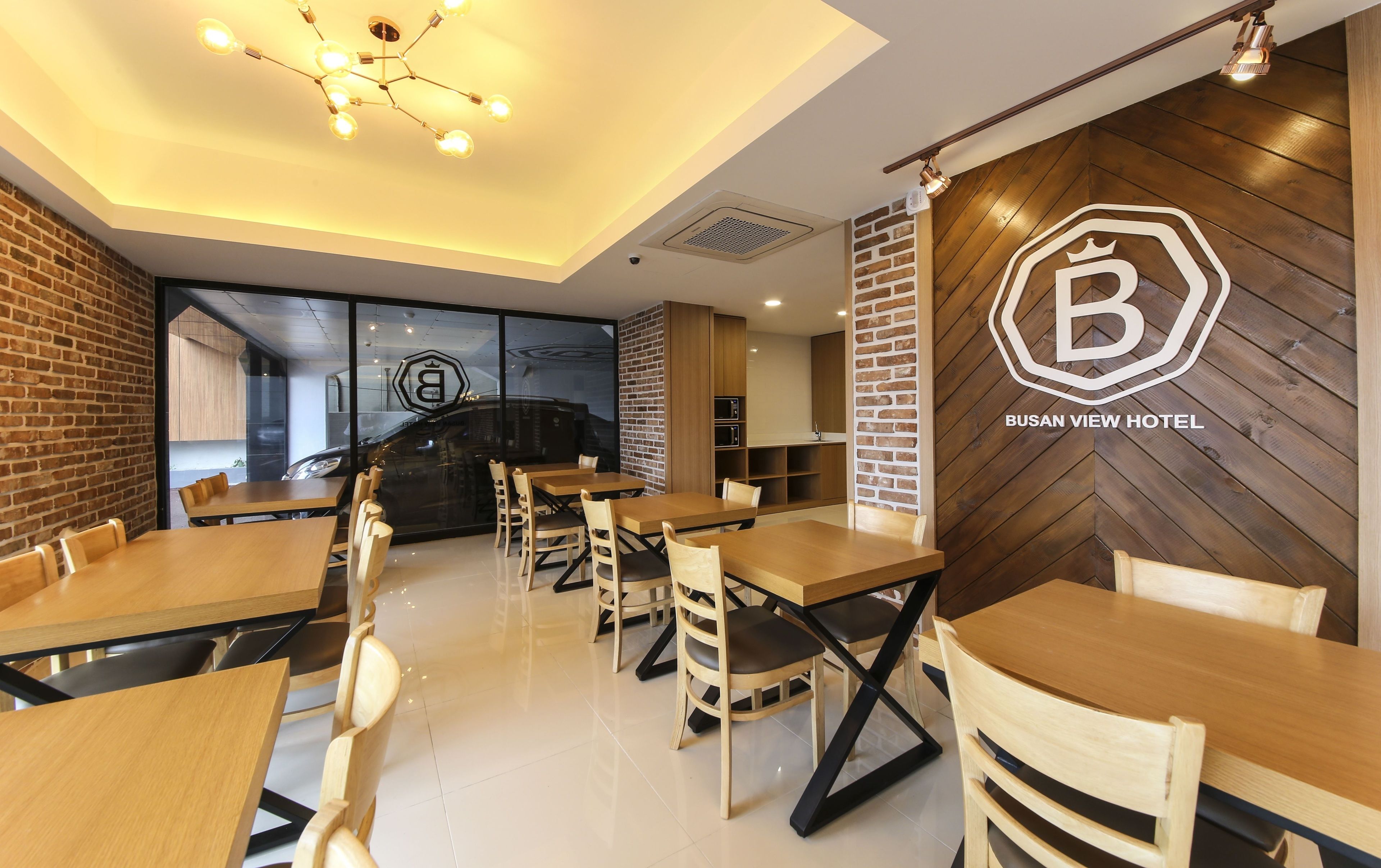 A modern hotel interior with wooden tables and chairs, a visible logo on the wall, and a kitchen area.