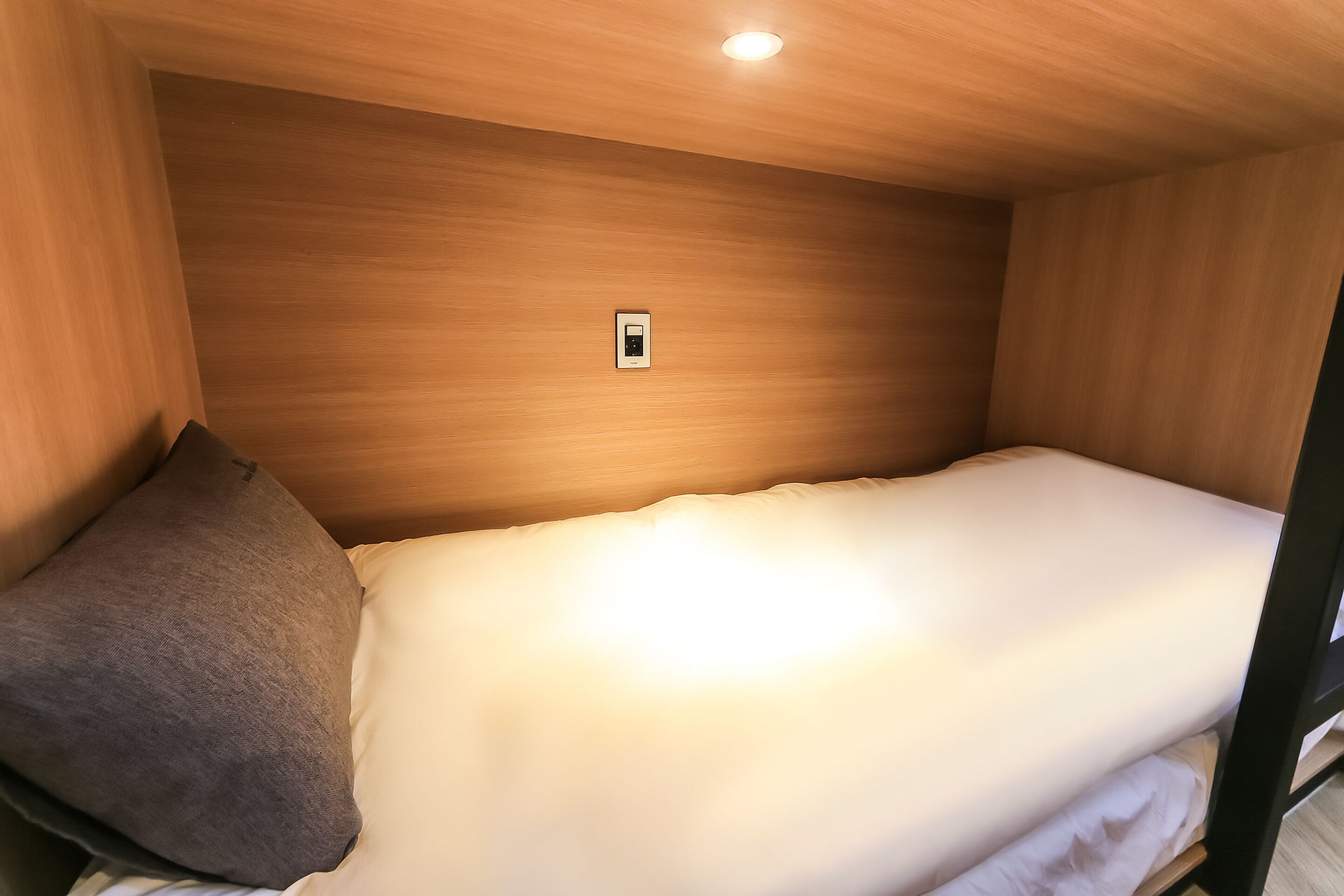 A single bed with a white mattress and a gray pillow in a room with wooden walls and a ceiling light.