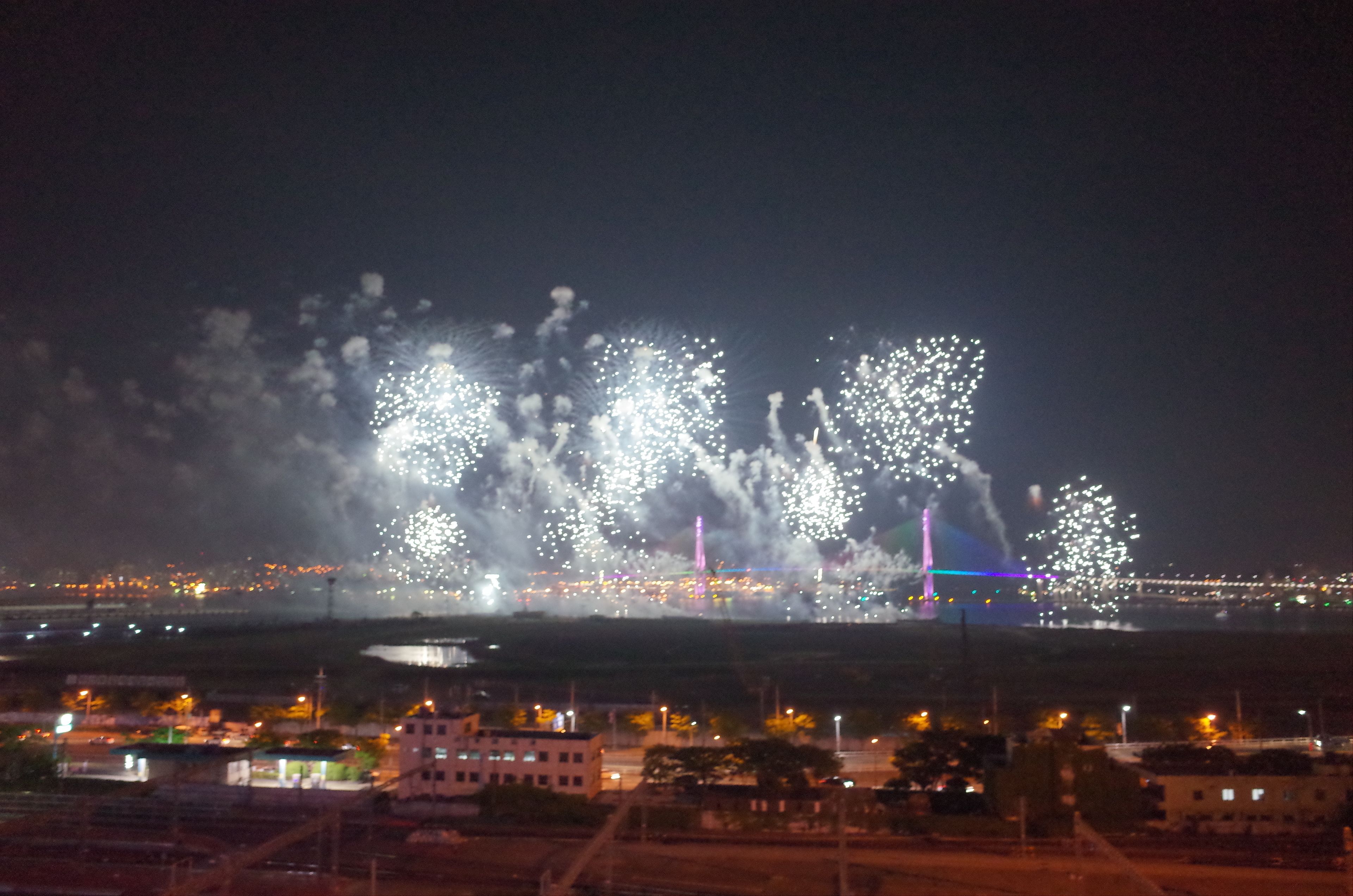 Une vue nocturne d’un feu d’artifice illuminant une ville, avec des bâtiments et un pont en arrière-plan.