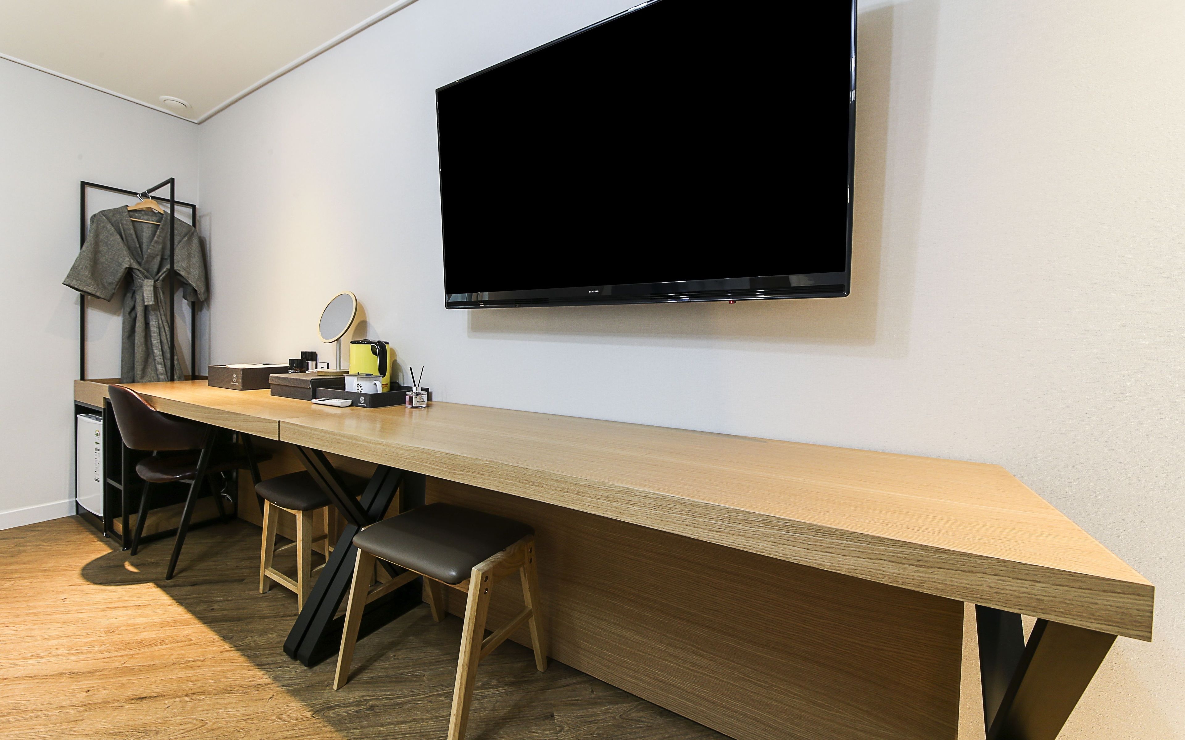 A modern room with a long wooden desk, two black chairs, a flat-screen TV mounted on the wall, and a coat rack with a grey coat.
