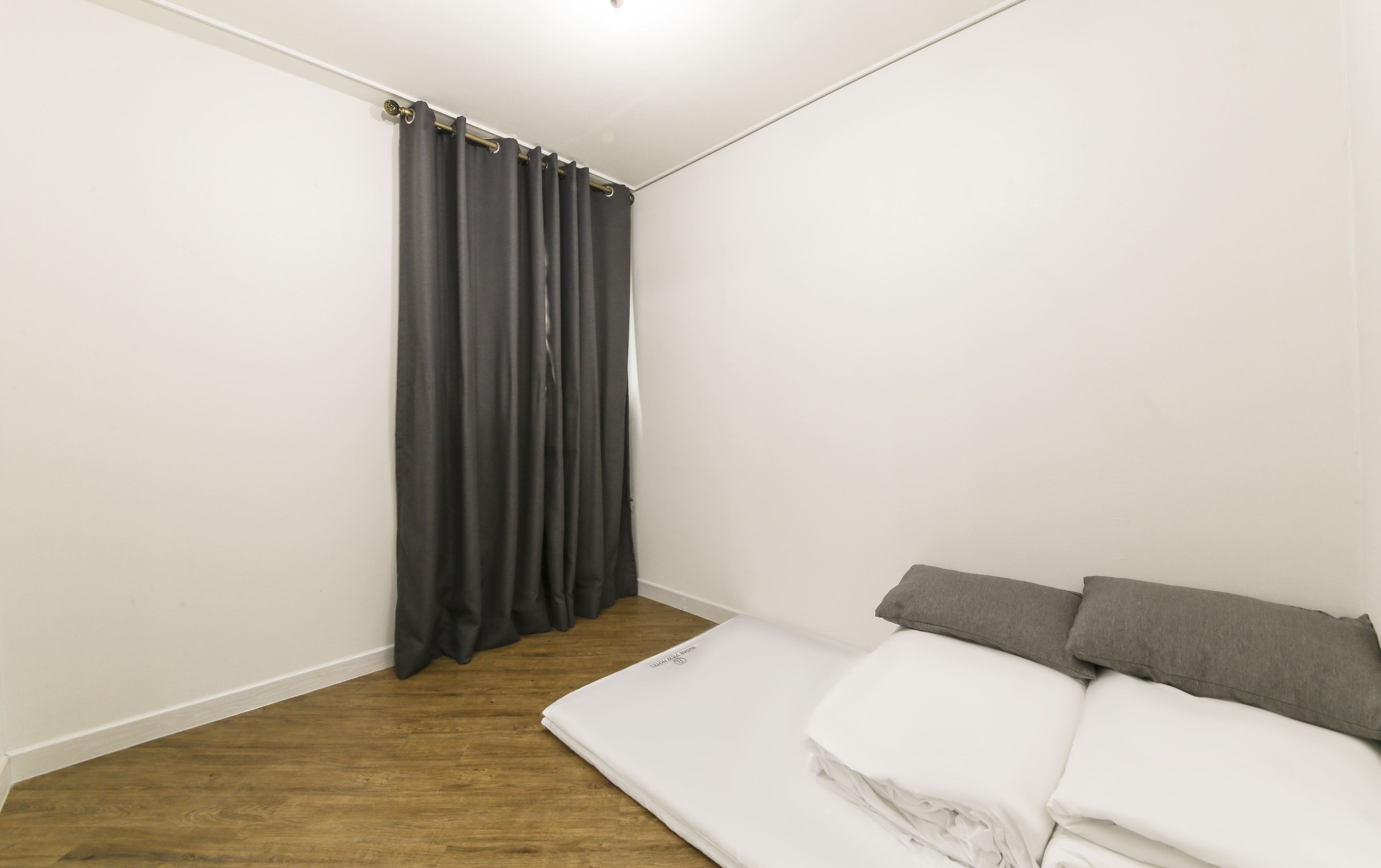 A bedroom with a wooden floor, a bed with white bedding and gray pillows, and a dark curtain on the window.