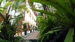 Un sentier bordé de végétation luxuriante, typique des jardins tropicaux, mène à un bâtiment doté d’un espace extérieur aménagé avec une zone de repos couverte.