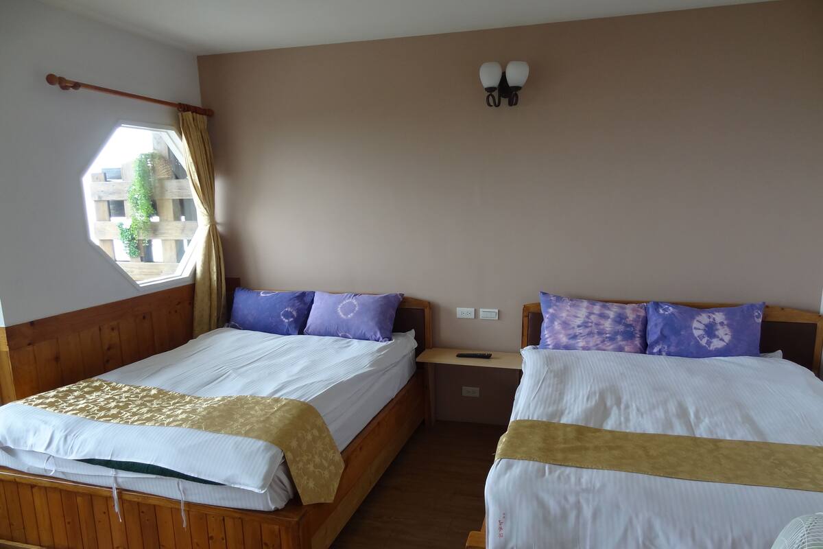 Una habitación con dos camas, cada una con ropa blanca de cama y almohadas moradas. Hay una ventana con cortina, una mesita de noche con control remoto y una lámpara fijada a la pared.