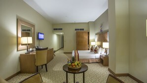 A hotel room with a bed, a desk, a chair, a small table with fruit, a television, and a wardrobe. - Rose View Hotel (Sylhet)