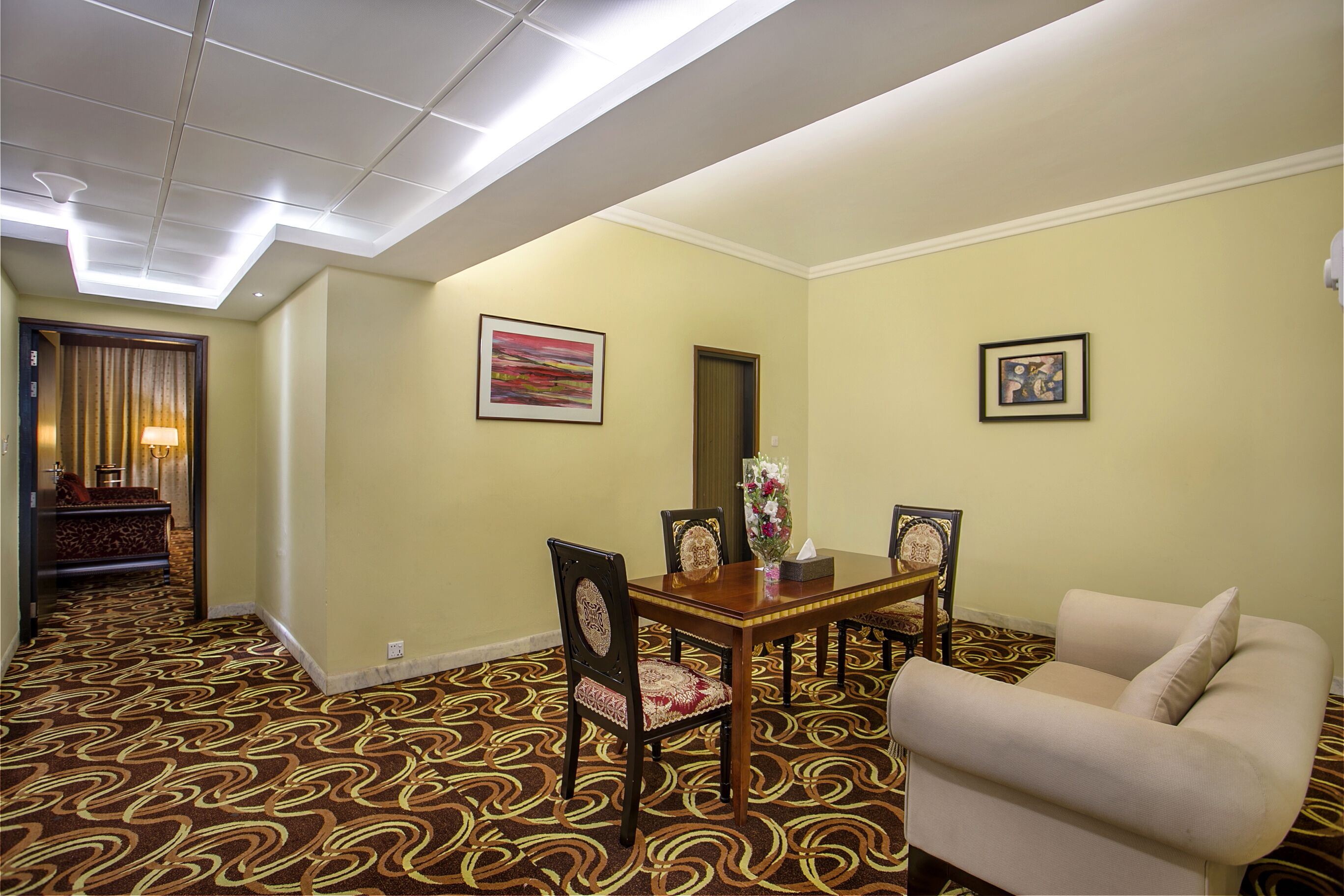 A hotel room with a dining table, chairs, a sofa, a patterned carpet, and two framed pictures on the walls.