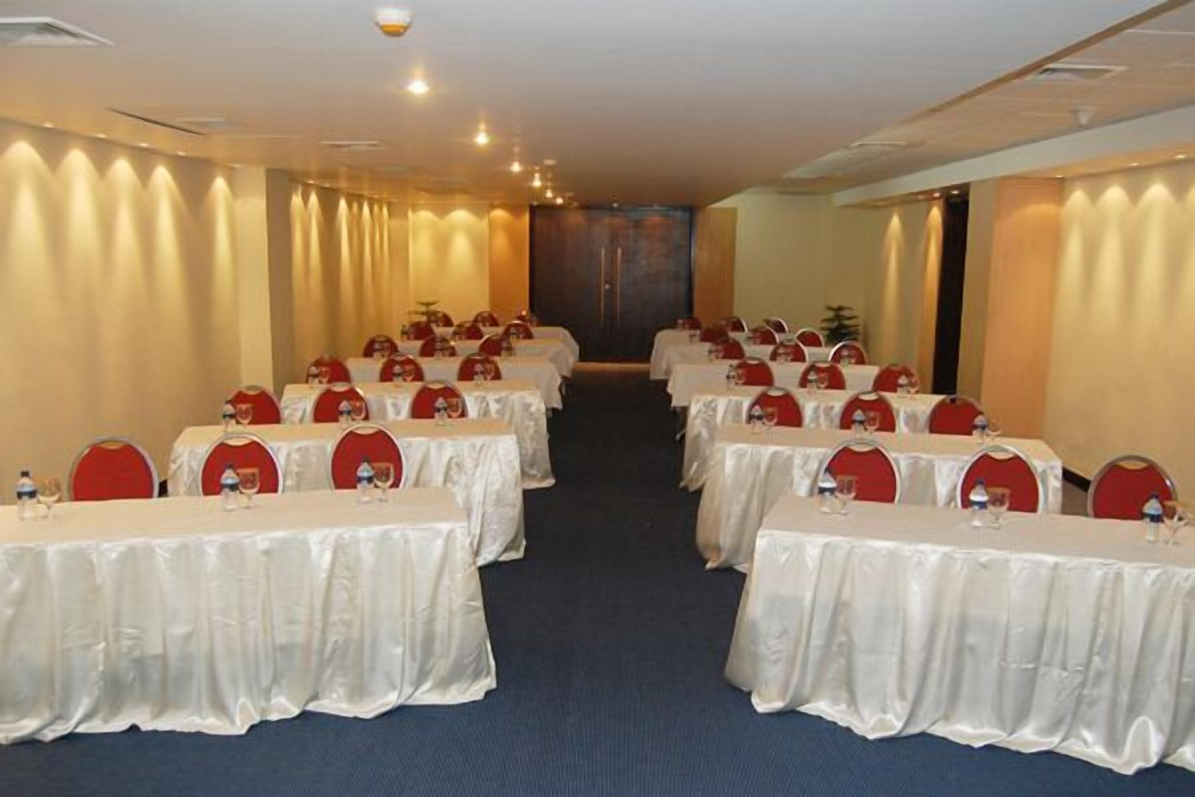 A conference room with rows of tables, each with a white tablecloth and red chairs.
