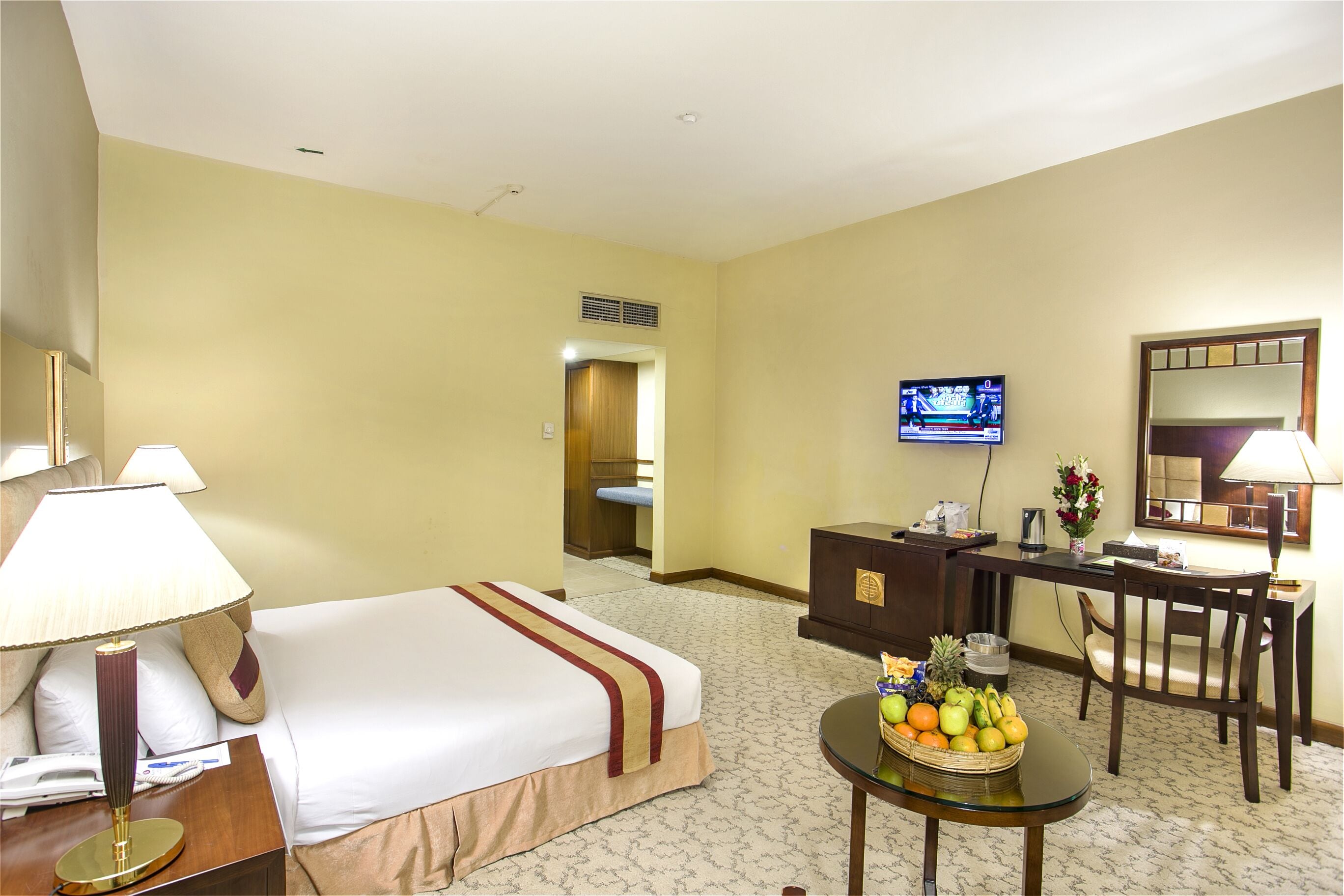 A hotel room with a bed, bedside table, lamp, desk, chair, TV, and a fruit basket.