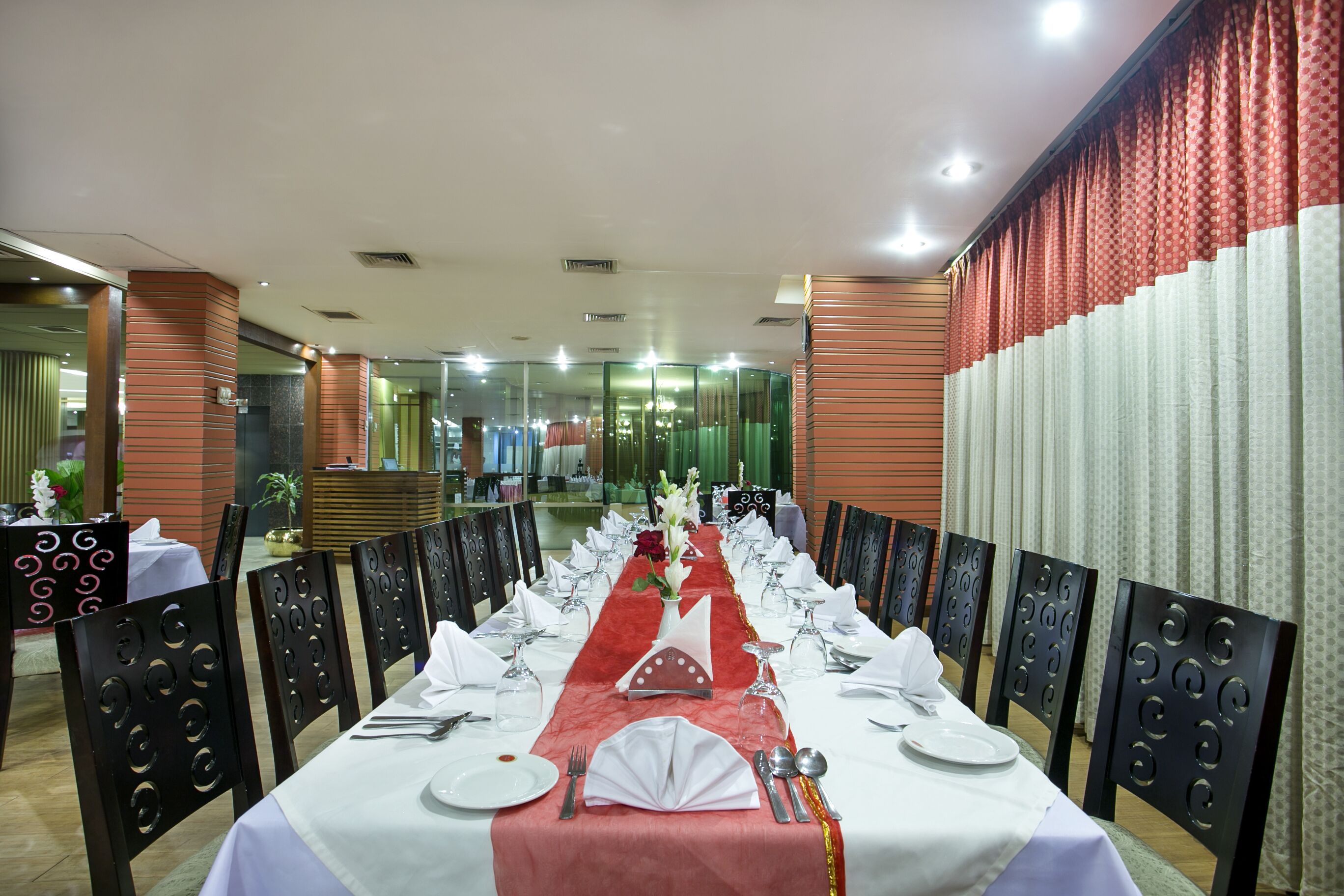 Mesa de jantar comprida posta para um evento formal, com toalhas brancas, arranjos vermelhos e cadeiras pretas com recortes circulares.