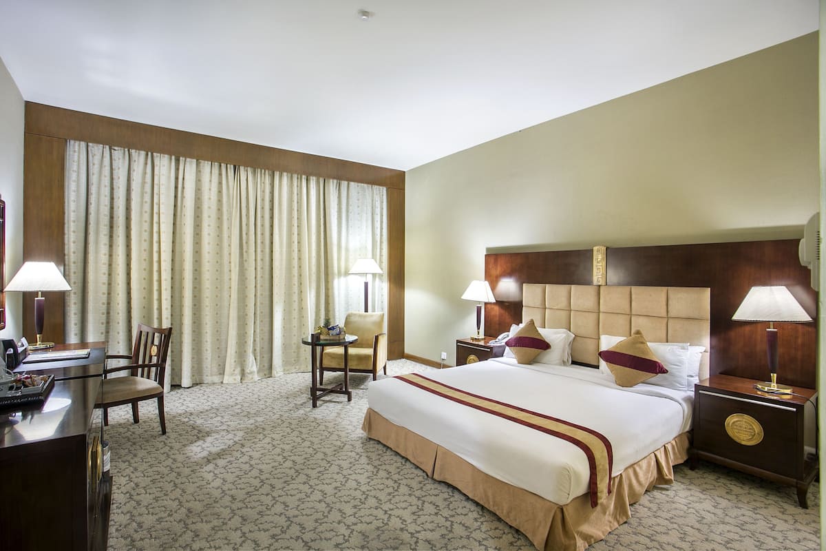 Une chambre d’hôtel avec un grand lit, deux tables de chevet avec des lampes, un bureau avec une chaise et une grande fenêtre avec des rideaux.