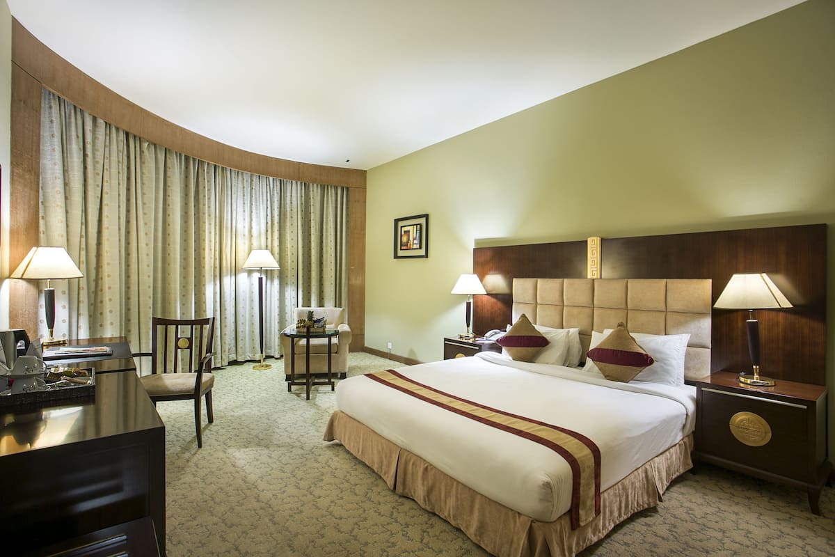 Une chambre d’hôtel avec un grand lit, deux tables de chevet avec des lampes, un bureau avec une chaise, une petite table et une grande fenêtre avec des rideaux.