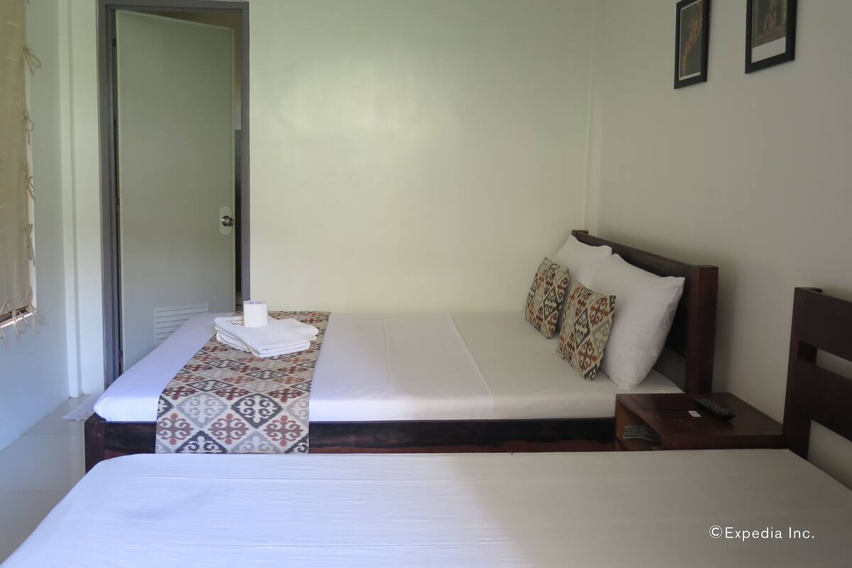 A hotel room with two beds, a bedside table with a remote control, patterned bedspreads, and a door.