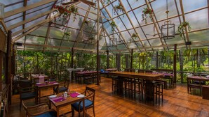 A greenhouse-style restaurant with wooden tables and chairs, hanging plants, and large windows offering a view of the surrounding greenery. - Munjoh Ocean Resort (Port Blair)
