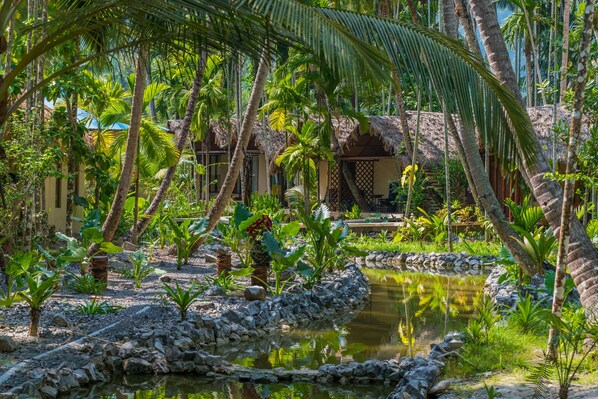 A tropical garden with a pond, palm trees, and a thatched-roof structure. - Munjoh Ocean Resort (Havelock Island)