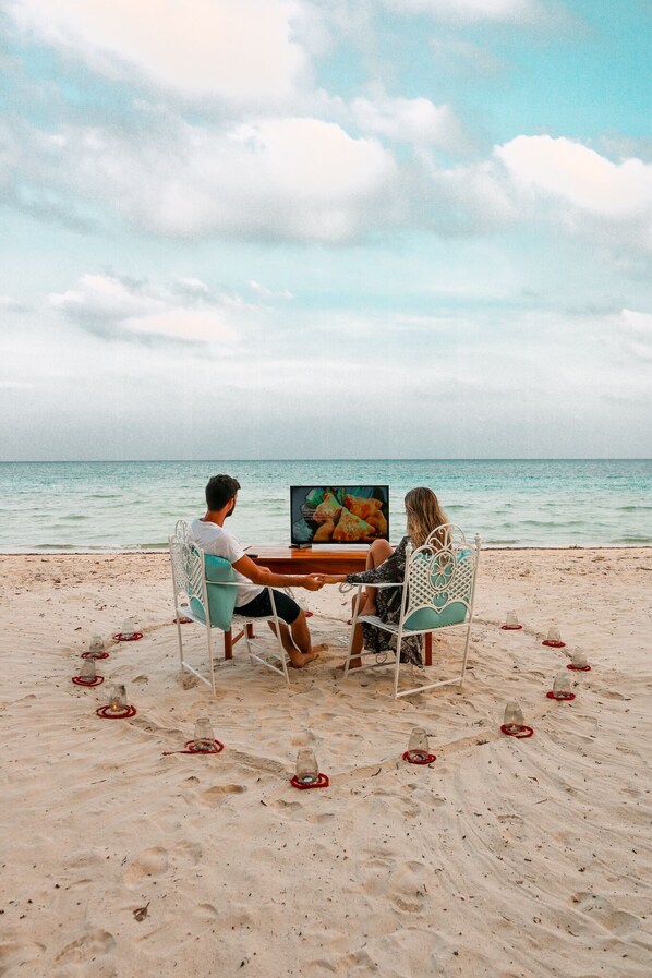 A beach with a table and chairs set up for two people, with a painting on the table. - Munjoh Ocean Resort (Havelock Island)