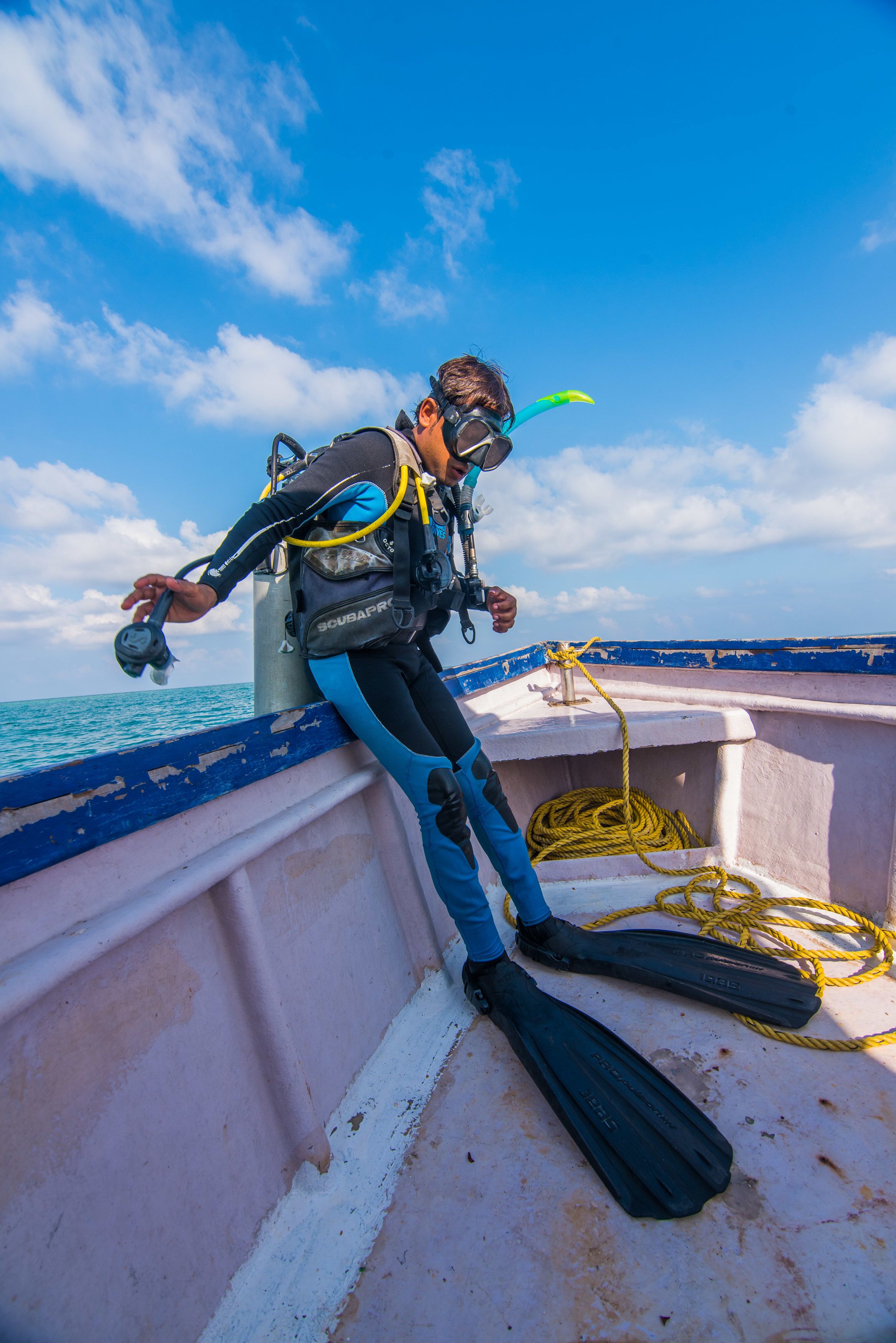 A diver with scuba gear leaning on a boat's edge, with the ocean and sky in the background.