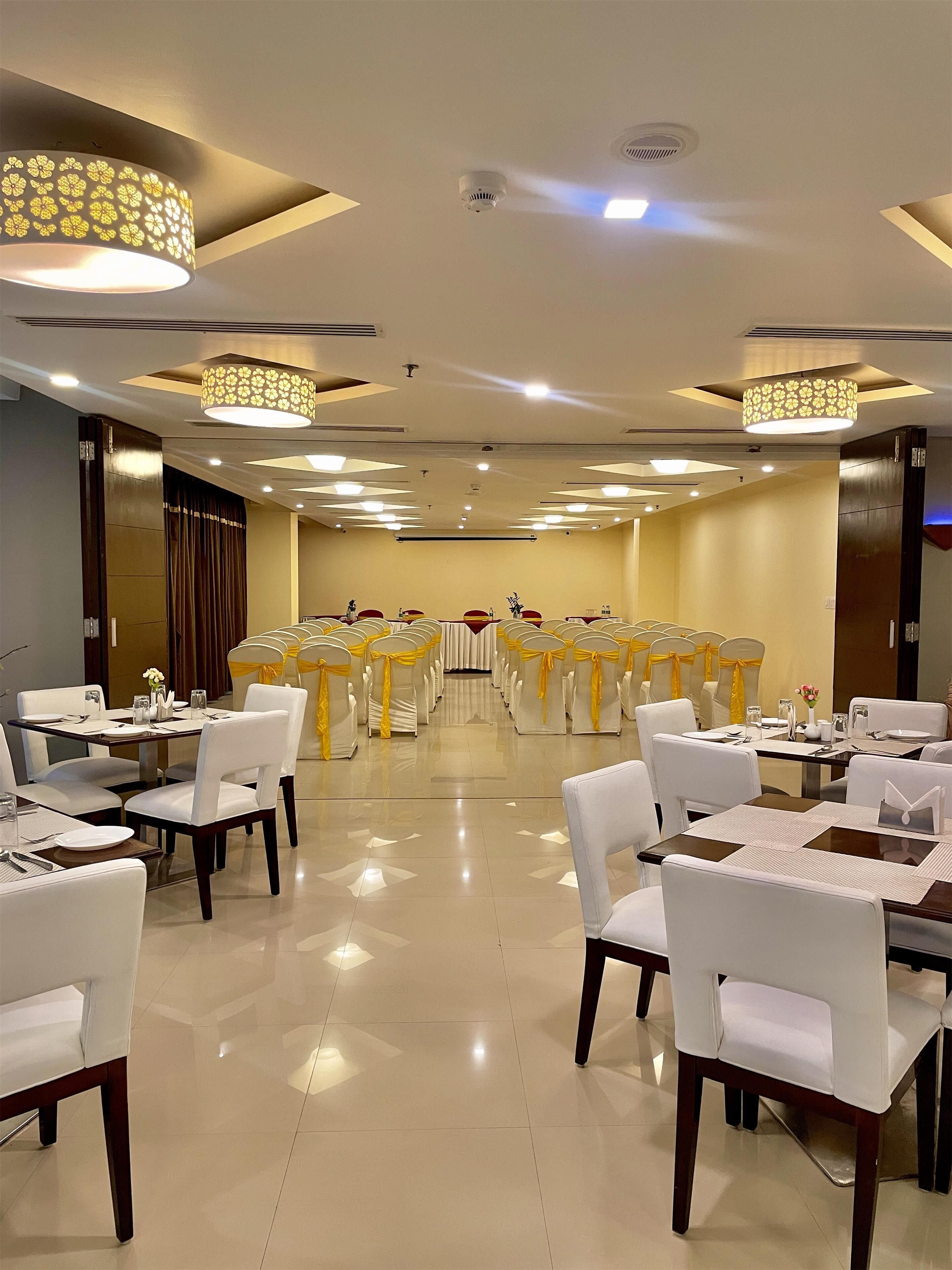 A banquet hall with rows of tables and chairs, ceiling lights, and a stage at the front.
