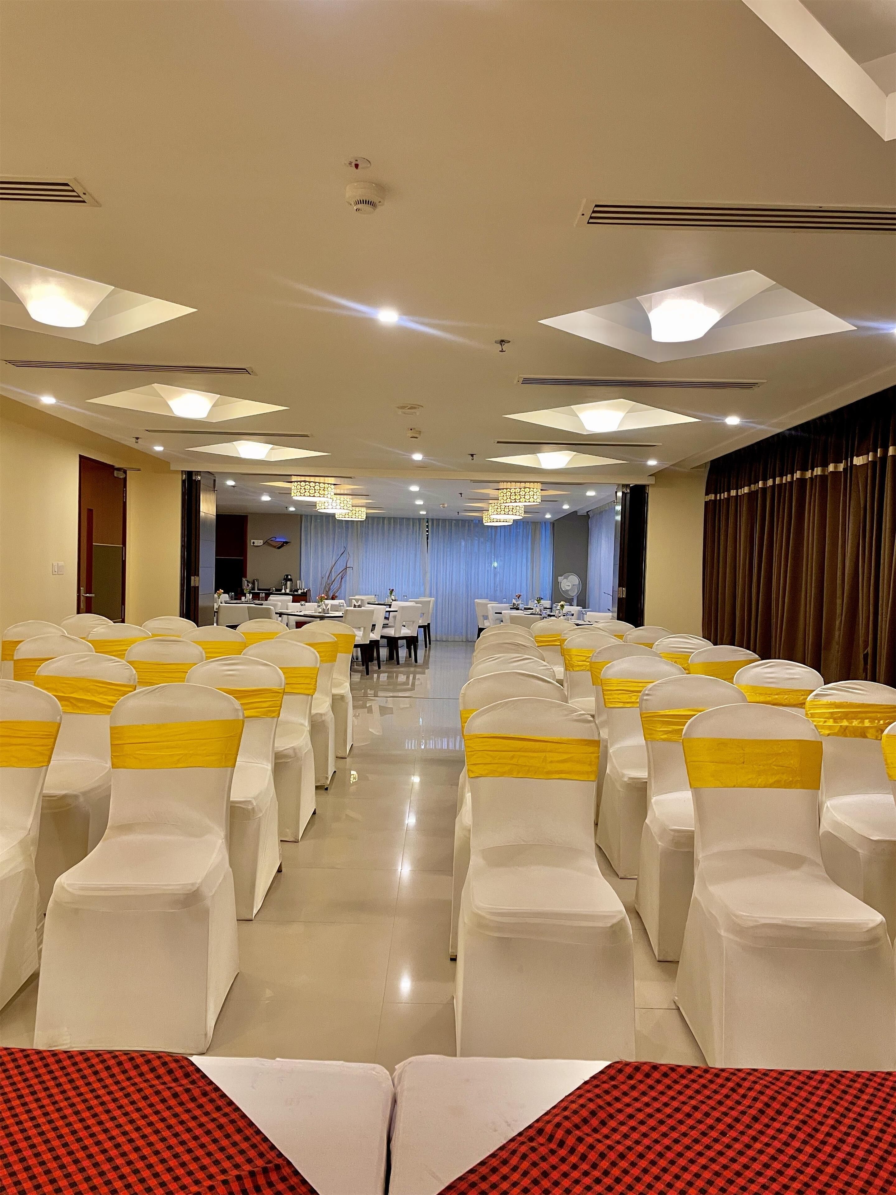 A banquet hall with rows of white chairs with yellow sashes, a stage area, and a ceiling with recessed lighting and air vents.