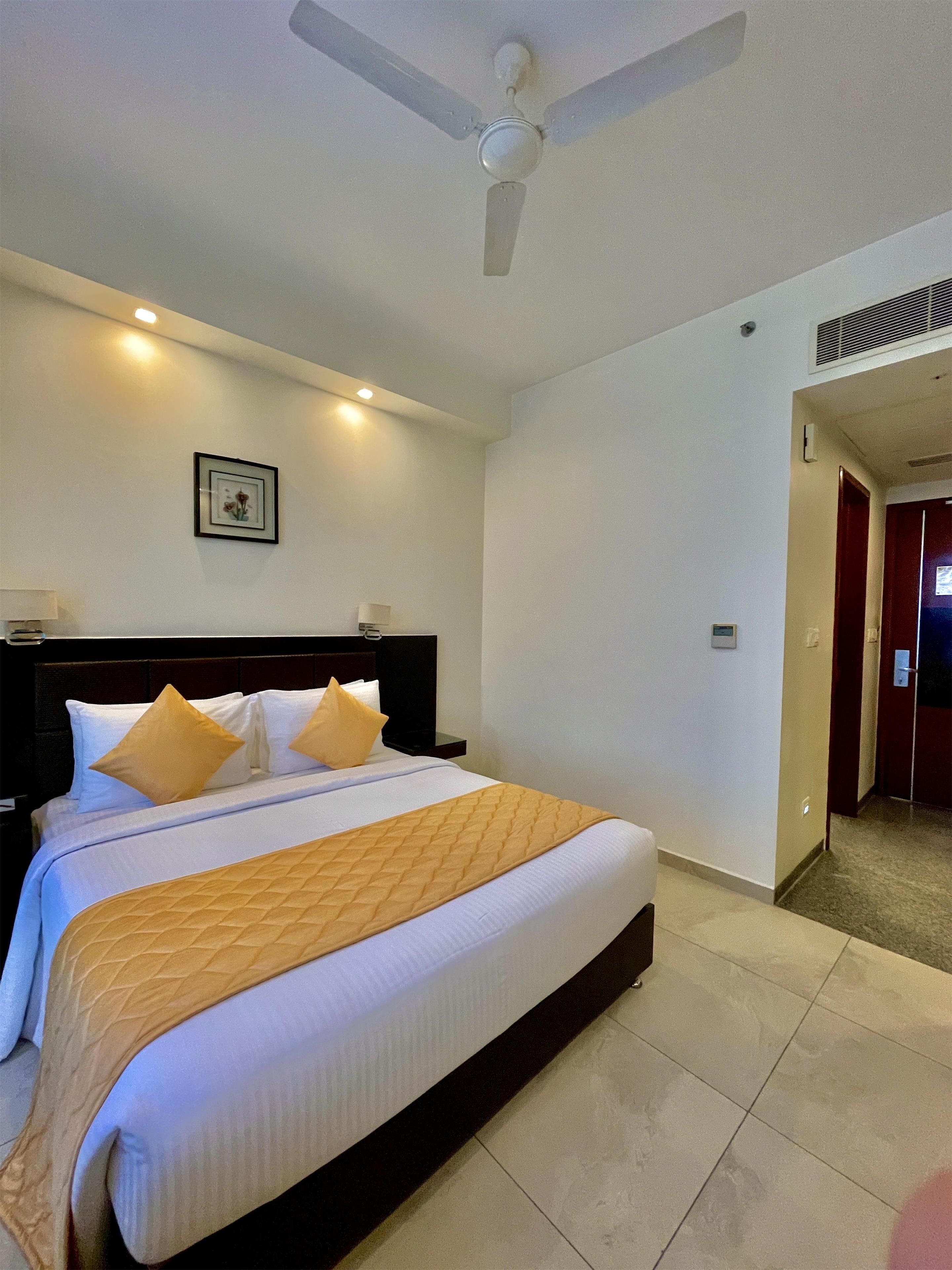 Quarto de hotel com uma cama grande, um ventilador de teto, um quadro pendurado na parede e uma porta que dá para outro cômodo.