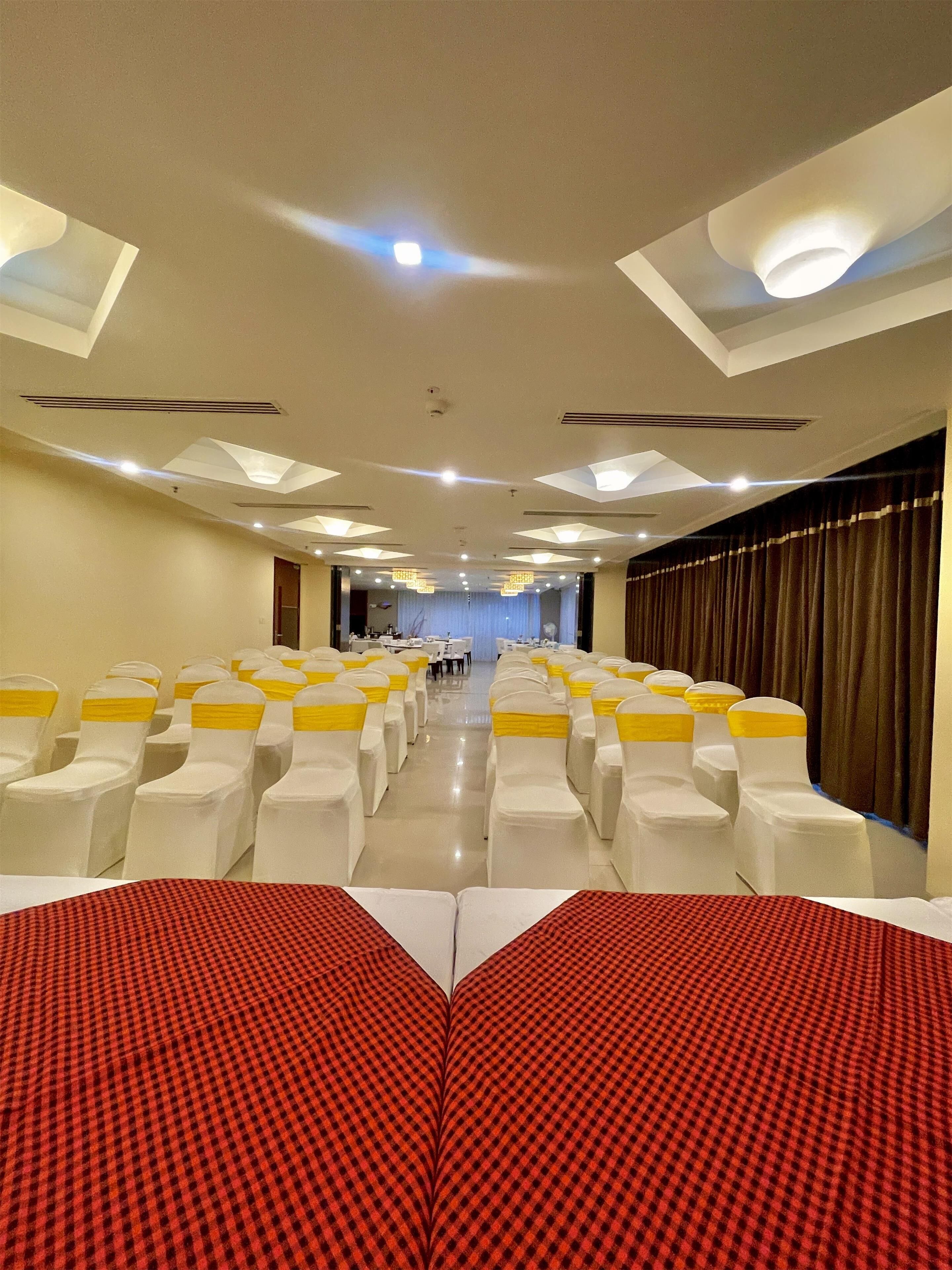 A hotel conference room with rows of white chairs with yellow sashes, a stage with a blue curtain, and a red checkered carpet in the foreground.