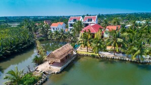 A riverside property with a thatched-roof structure, a pool, and several multi-story buildings with red roofs. - Lakeview Villa Homestay Hoi An (Da Nang)