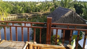 A wooden deck with a railing overlooking a body of water, surrounded by lush greenery and a thatched-roof structure. - Lakeview Villa Homestay Hoi An (Da Nang)