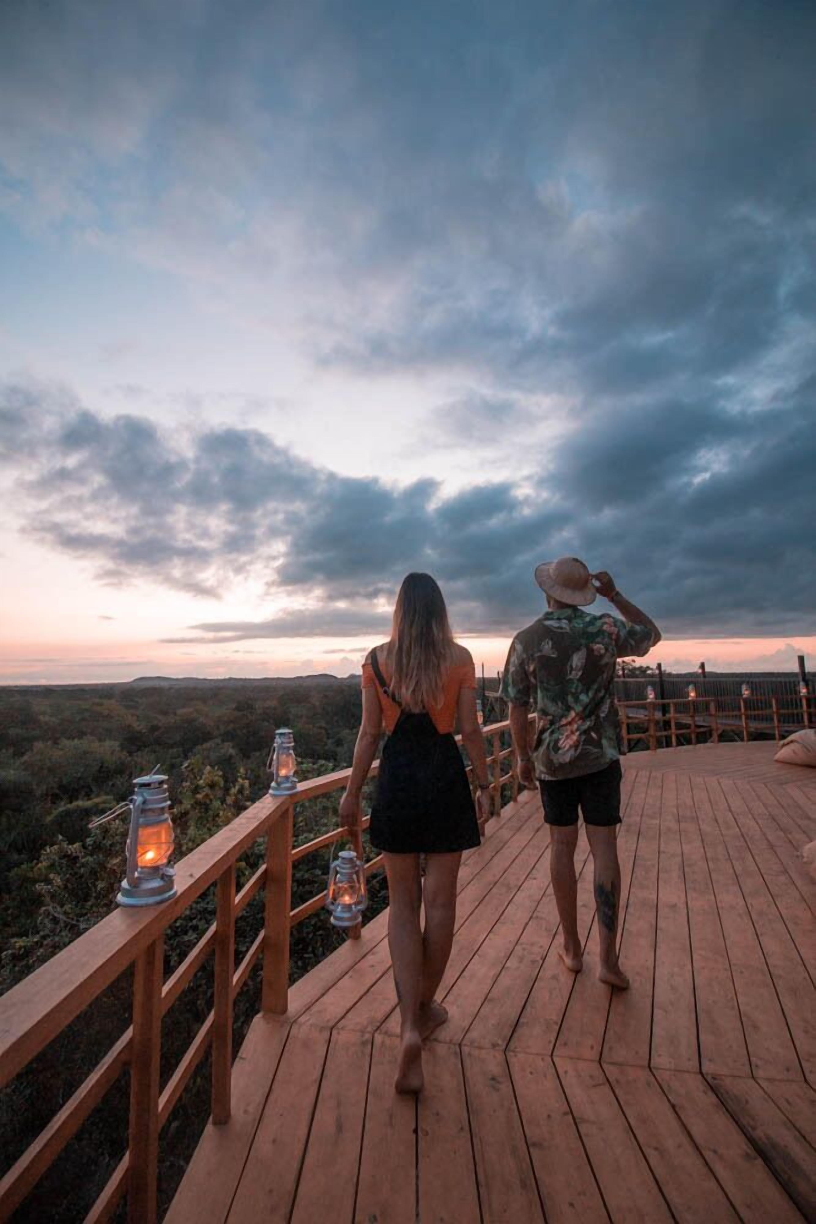 A couple walking on a wooden deck with lanterns, overlooking a landscape.
