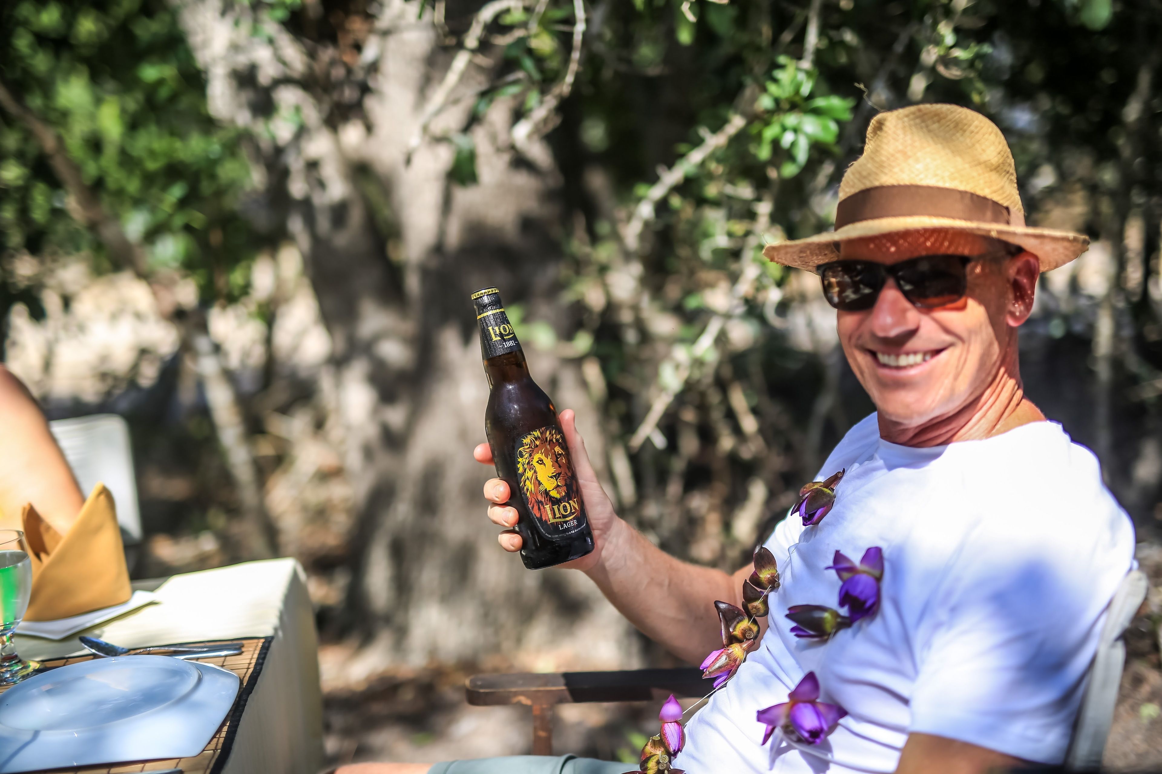 A person holding a beer bottle with a lion label, sitting outdoors with a table set for a meal.