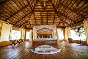 A room with a wooden floor, a bed with a canopy, a bedside table, a fan, and a window with curtains.