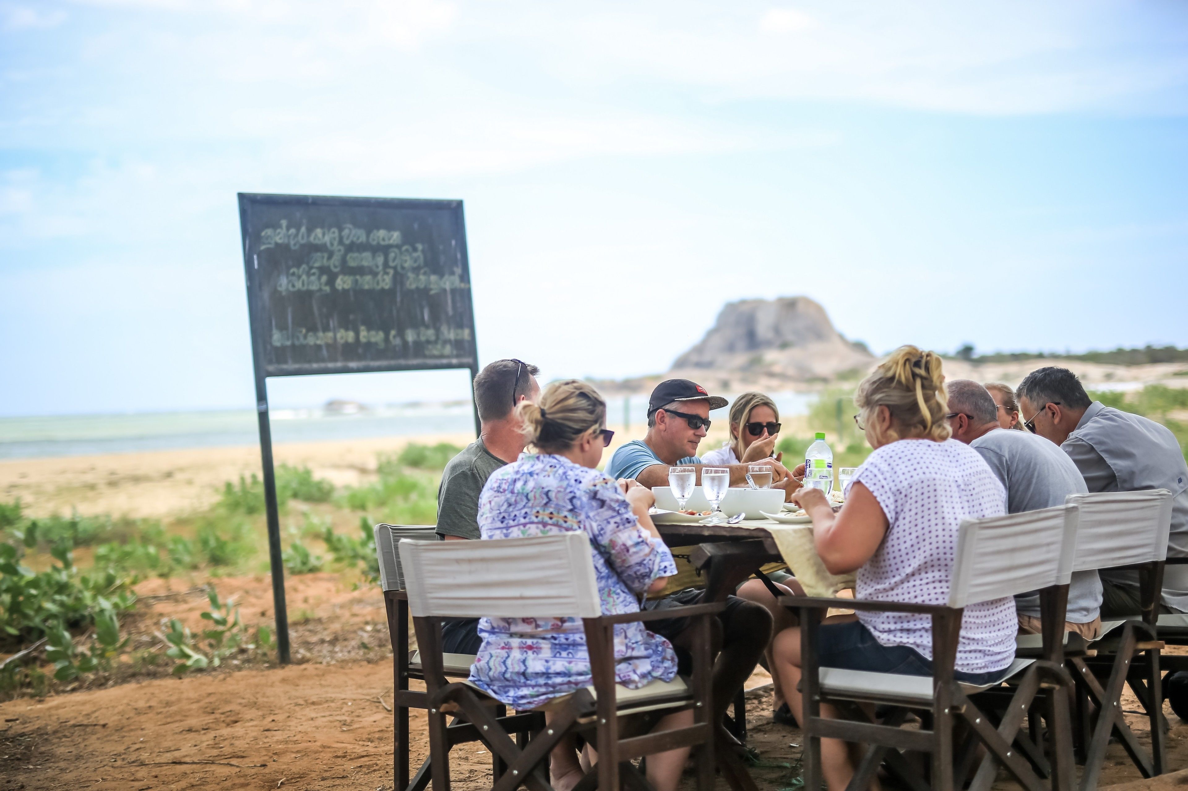 Un grupo de personas cenando en una mesa al aire libre con vistas a la playa y un cartelón al fondo.