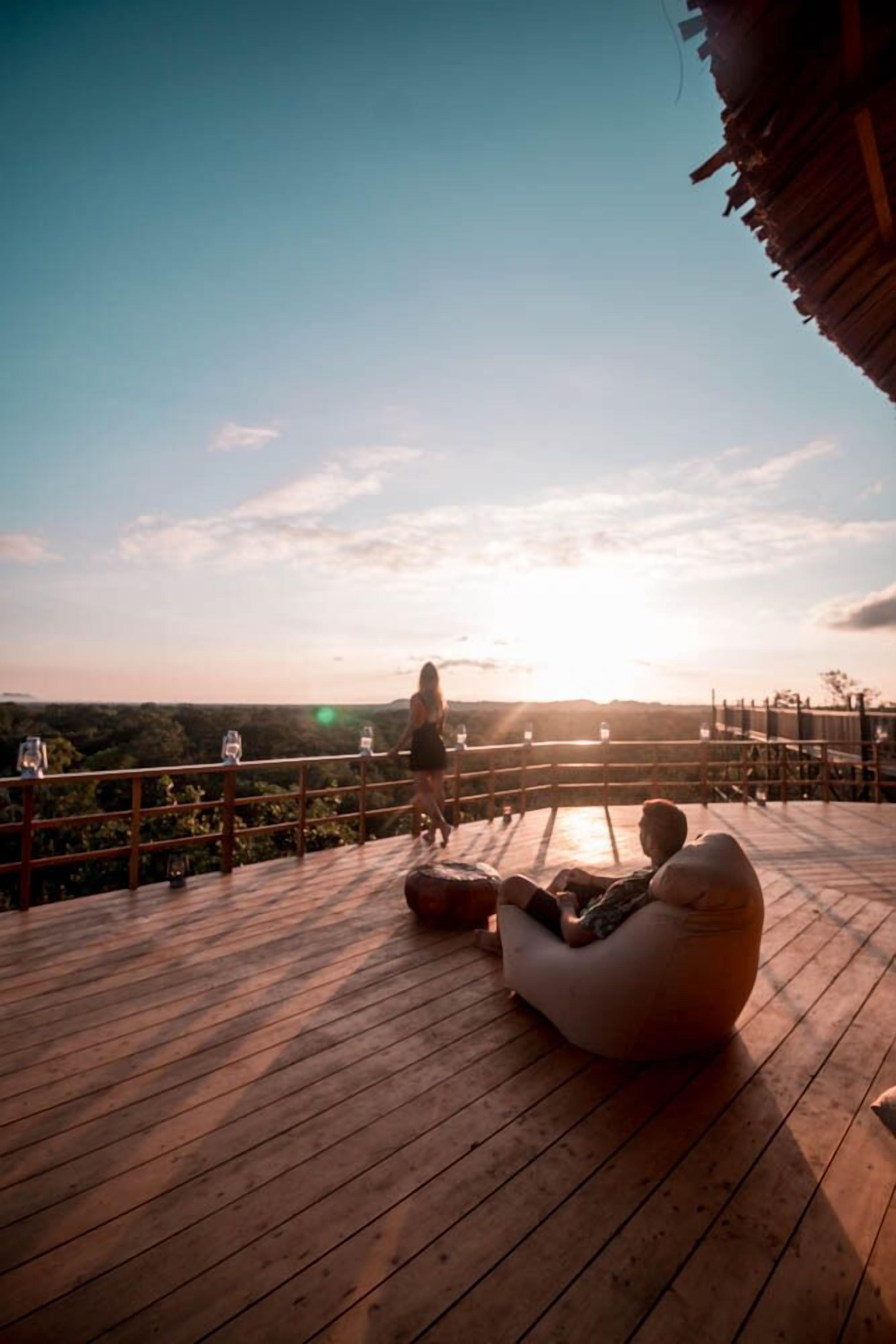 A wooden deck with a railing, a person walking, and a sunset in the background.