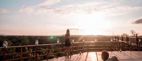 A wooden deck with a railing, a person walking, and a sunset in the background.