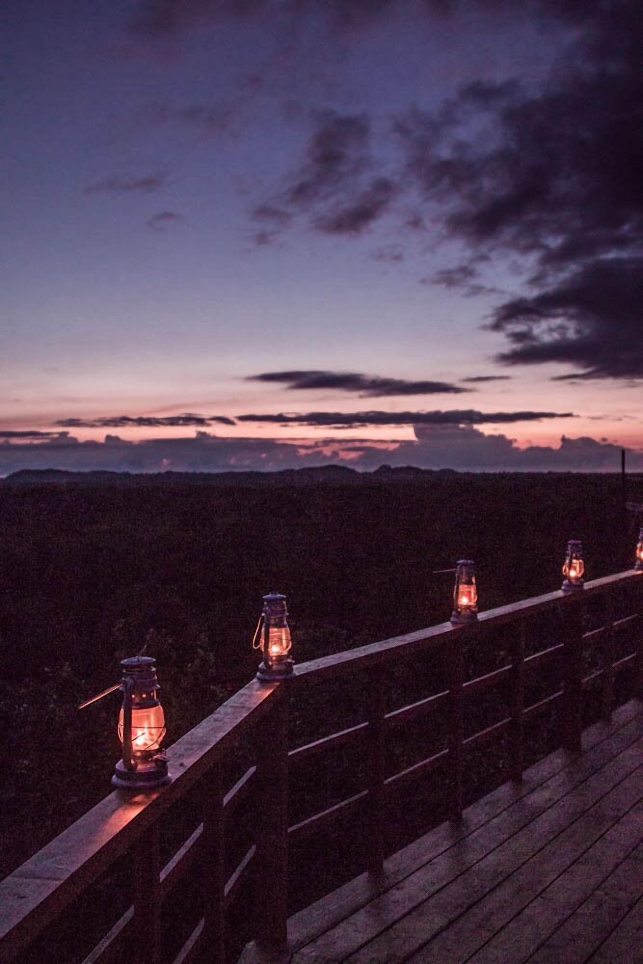 A wooden deck with lanterns along the railing, overlooking a mountainous landscape at dusk.