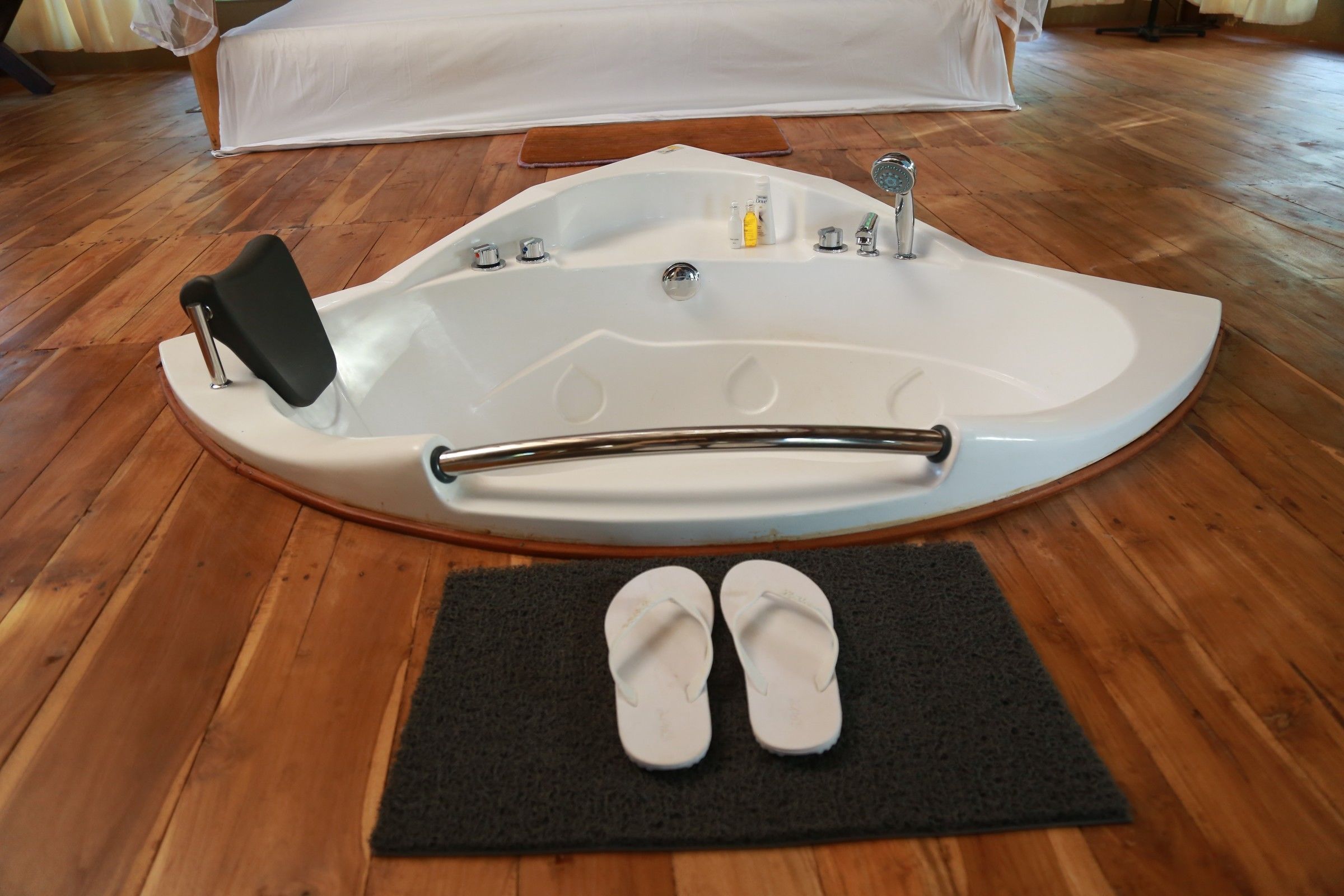 A large, freestanding bathtub with a built-in seat and a grab bar, placed on a wooden floor with a black mat in front.