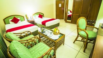 Habitación de hotel con dos camas individuales, una butaca verde acolchada, una silla de mimbre y una mesita con un cesto de plátanos.