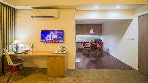 A hotel room with a wooden desk, a television displaying a news channel, a kitchenette, and a dining area with chairs and a table. - Asia Pacific Hotel (Dhaka)