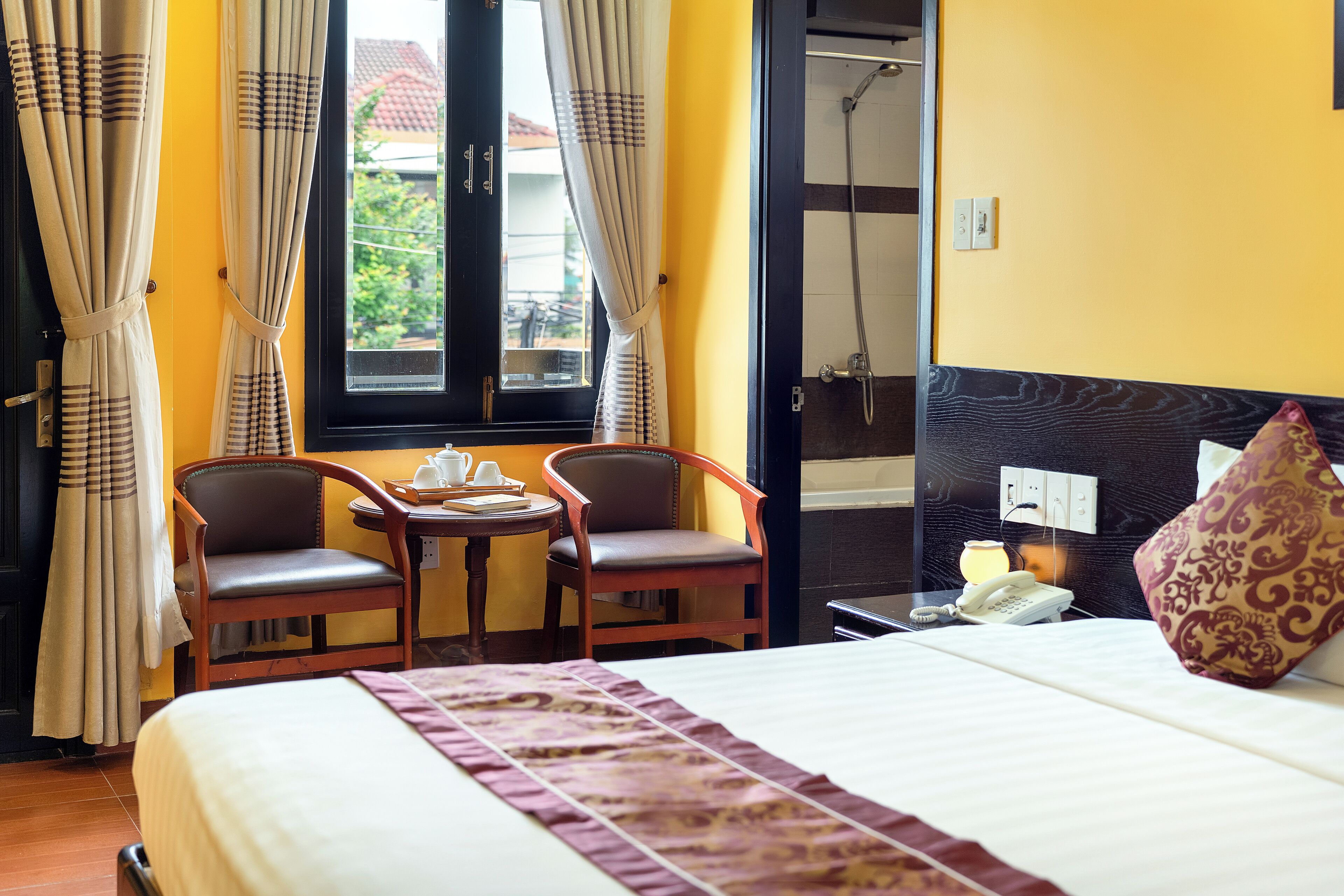A hotel room with a bed, two chairs, a small table with a teapot, a telephone, and a window with curtains.
