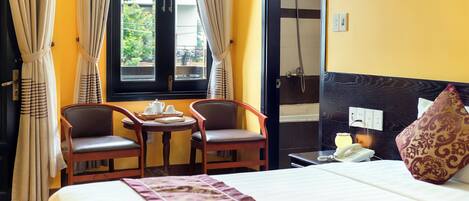 A hotel room with a bed, two chairs, a small table with a teapot, a telephone, and a window with curtains.