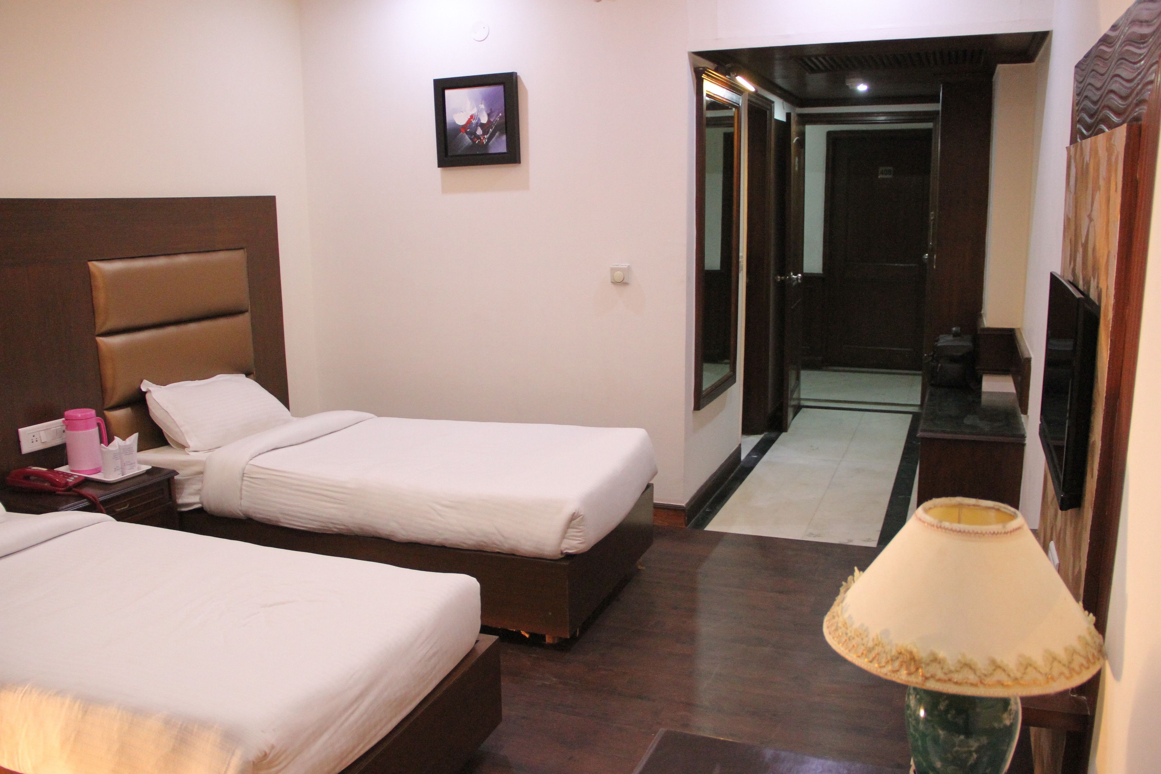 Habitación de hotel con dos camas, una mesita de noche con lámpara, un cuadro en la pared y una puerta que da a un pasillo.