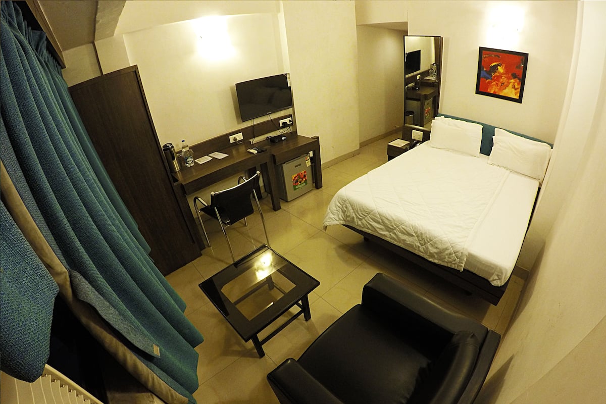 Une chambre d’hôtel avec un lit, un bureau avec une télévision, une chaise, une petite table et une fenêtre avec des rideaux.