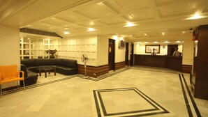 A spacious lobby with a checkered floor, a black leather sofa, an orange chair, and a fan. - Tex-Palazzo Hotel (Surat)