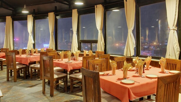 A restaurant with tables set for dining, large windows with curtains, and a city view at night. - Tex-Palazzo Hotel (Surat)
