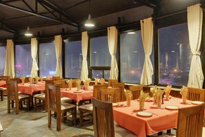 A restaurant with tables set for dining, large windows with curtains, and a city view at night. - Tex-Palazzo Hotel (Surat)