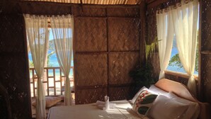 A bed with a white mattress and pillows, a wooden headboard, and a view of the beach through a window with white curtains.