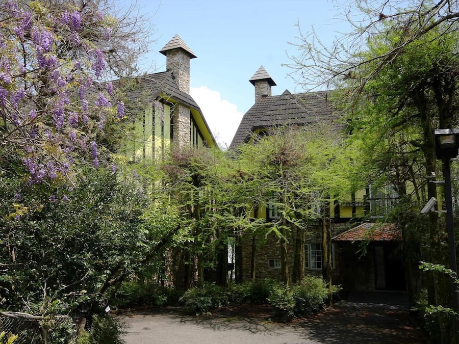 A traditional stone building with a steeply pitched roof, surrounded by lush greenery and purple flowering trees.