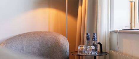 A hotel room with a grey armchair, a small table with bottles, a floor lamp, and a radiator.