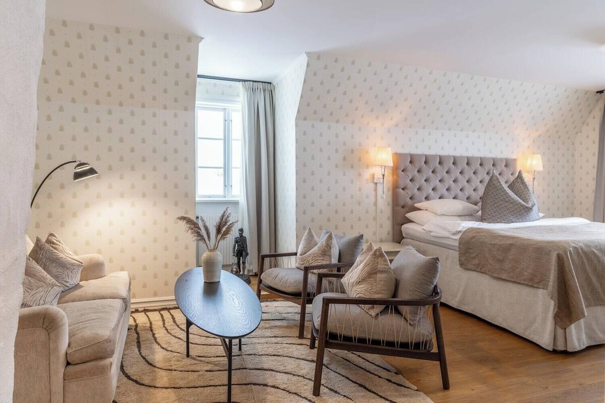 A hotel room with a bed, two chairs, a small table, a lamp, and a window with curtains.