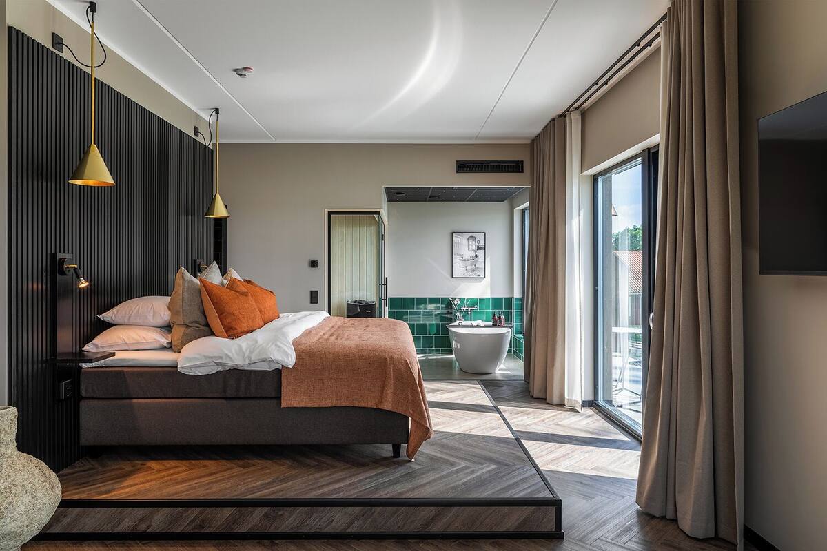 A modern hotel room with a bed, bedside lamps, a bathroom with a bathtub, and a large window with curtains.